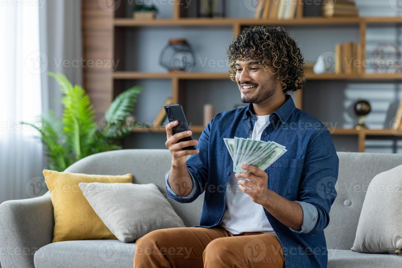 Happy man checking bank account balance using a mobile app and holding a  fan of cash, sitting on the couch at home. 62293377 Stock Photo at