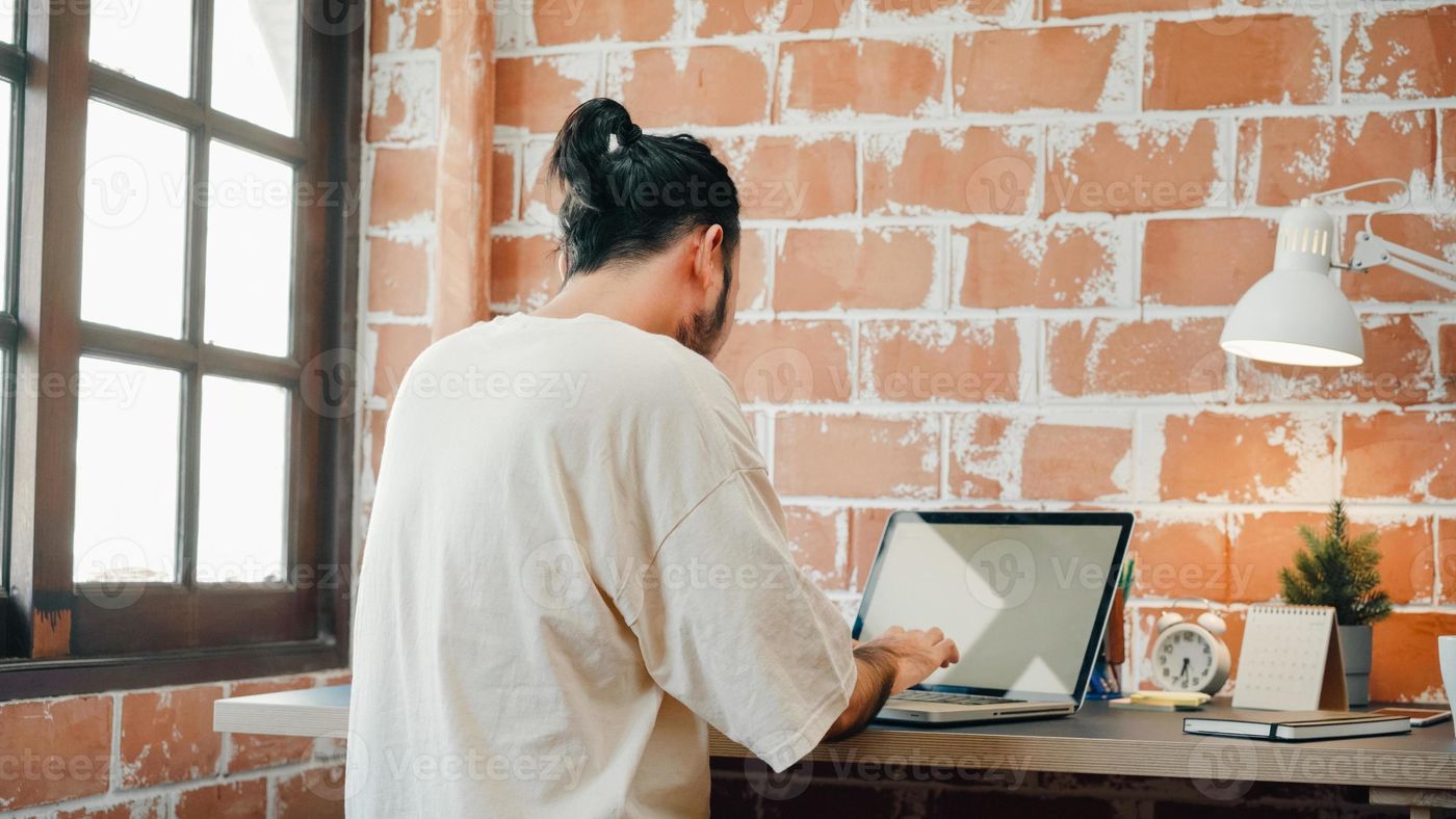 Back view freelance hipster modern Asian man designer working home using laptop in living room at home. Happy young male work while sitting